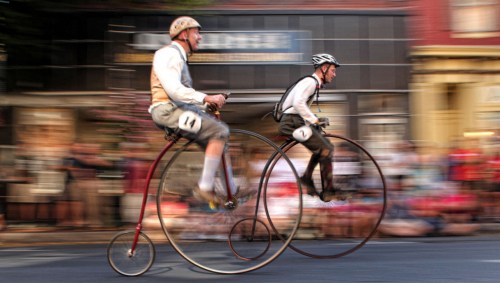 Two men in vintage attire race on tall, old-fashioned bicycles, with a blurred crowd in the background.