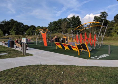A colorful outdoor playground with climbing structures and fitness equipment, surrounded by grass and trees.