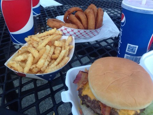 A burger with cheese and bacon, crinkle-cut fries, onion rings, and two cups of Pepsi on a table.