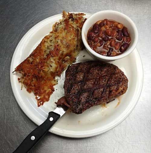 A plate with a grilled steak, crispy hash browns, and a bowl of baked beans, with a knife beside the steak.