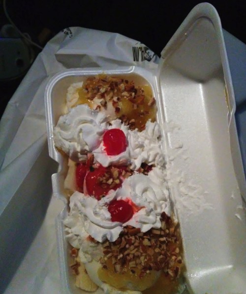 A dessert in a takeout container featuring bananas, whipped cream, cherries, and chopped nuts.