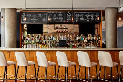 A stylish bar with a wooden counter, high stools, and a large selection of drinks displayed on chalkboards above.