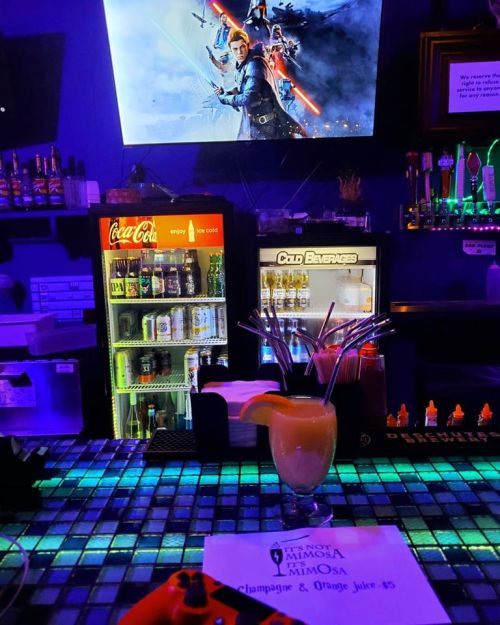 A colorful bar scene featuring a cocktail, gaming controller, and a screen displaying a fantasy-themed image.