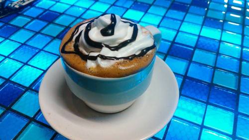 A cup of coffee topped with whipped cream and chocolate drizzle, sitting on a white saucer against a blue tiled background.