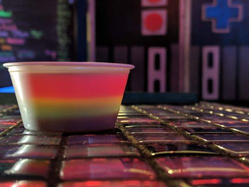 A colorful rainbow dessert in a clear cup sits on a reflective surface with vibrant lighting in the background.