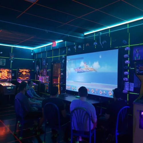 A dimly lit arcade with neon accents, featuring a group of people watching a video game on a large screen.