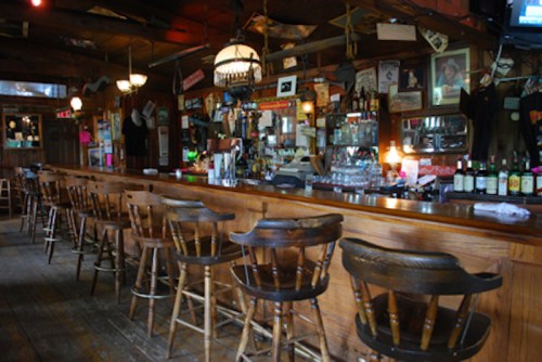 A rustic bar interior with wooden stools, a long counter, and shelves filled with various bottles and memorabilia.