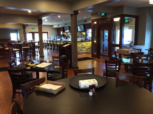 Interior of a restaurant featuring wooden tables, chairs, and a bar area with a stone counter.