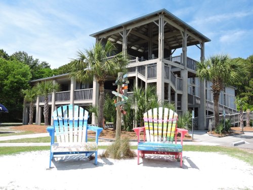Two colorful adirondack chairs in front of a beach house, surrounded by palm trees and a sandy pathway.