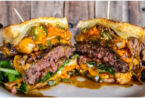 A close-up of a juicy burger with melted cheese, toppings, and toasted bread, served on a rustic wooden background.