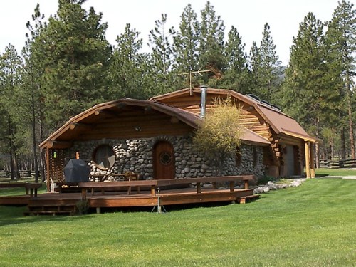 A rustic stone and wood cabin surrounded by tall pine trees and a green lawn, featuring a large deck and unique architecture.