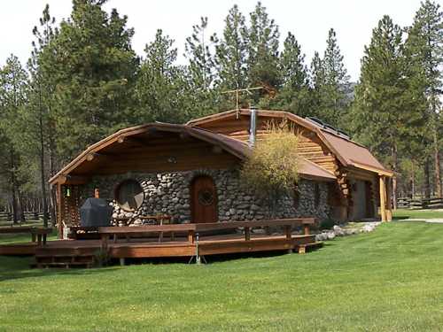 A rustic stone and wood cabin surrounded by tall pine trees and a green lawn, featuring a large deck and unique architecture.