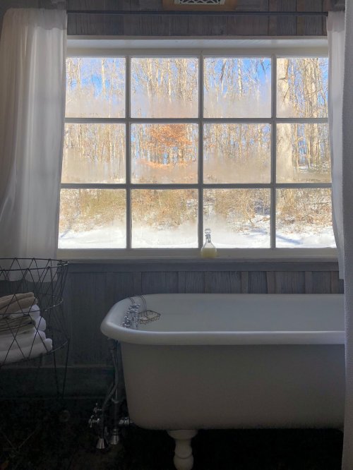A cozy bathroom with a vintage tub, featuring a foggy window overlooking a snowy landscape and trees.