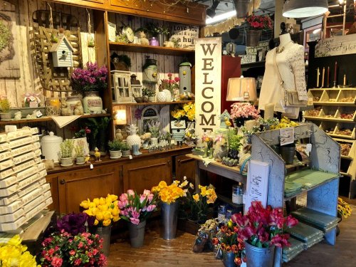 A cozy shop filled with flowers, plants, and decorative items, featuring a large "WELCOME" sign and charming displays.
