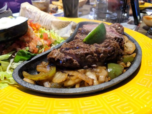 Sizzling steak fajitas on a hot plate, served with grilled onions, peppers, and a lime wedge.