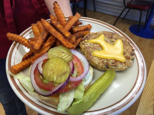 A plate with a veggie burger topped with cheese, lettuce, tomato, onion, pickles, and a side of sweet potato fries.