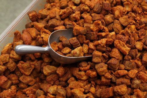 A close-up of crispy, golden-brown pork cracklings in a container, with a metal scoop resting on top.