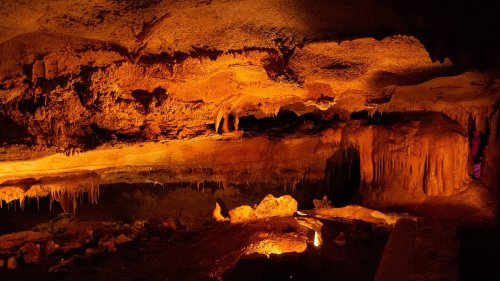 A dimly lit cave interior with orange hues, featuring stalactites, stalagmites, and a reflective water surface.