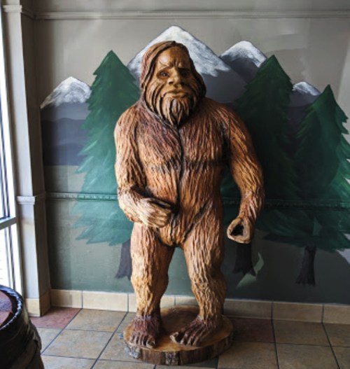 A wooden statue of Bigfoot stands against a mural of mountains and trees, showcasing intricate carvings and details.