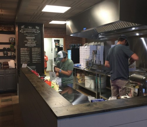 Two people working in a donut shop kitchen, preparing ingredients and cooking behind a counter.