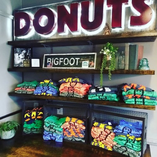 A display of colorful t-shirts stacked on shelves, with a neon "DONUTS" sign and a "RIGFOOT" framed picture above.