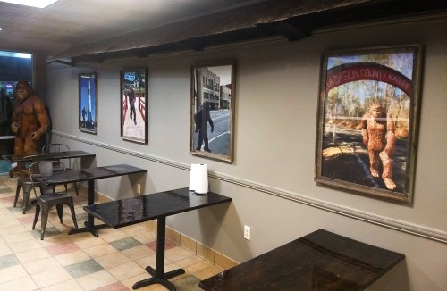 A cozy restaurant interior featuring wooden tables, metal chairs, and framed artwork, including a Bigfoot statue.