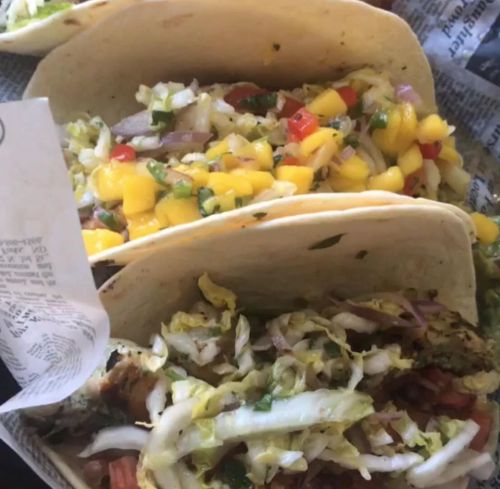 Three tacos filled with fresh vegetables, mango salsa, and shredded lettuce, served in a paper-lined basket.