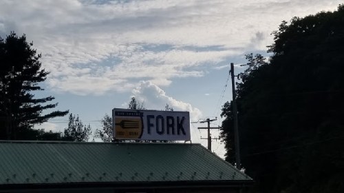 Sign for "The Fork" restaurant against a cloudy sky, with trees and power lines in the background.