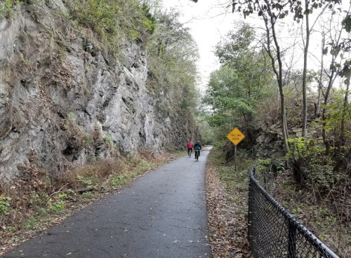 Two cyclists ride along a paved path surrounded by rocky cliffs and greenery, with a warning sign nearby.