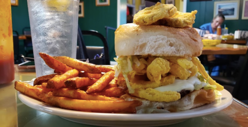 A plate with a stacked burger topped with lettuce and fried onions, served with sweet potato fries and a glass of water.