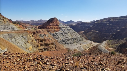 A vast, colorful mining landscape with steep, layered hills and a clear blue sky in the background.