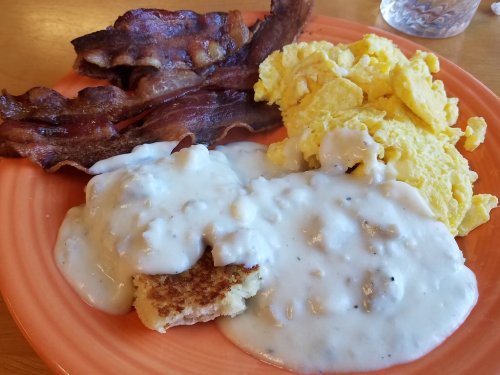 A plate of breakfast featuring crispy bacon, scrambled eggs, and biscuits smothered in creamy gravy.
