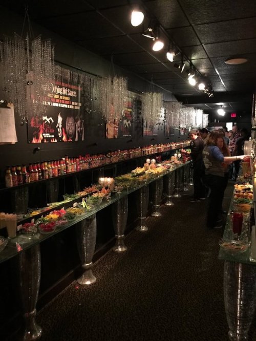 A dimly lit bar with a long table displaying various sauces, garnishes, and drinks, surrounded by decorative elements.