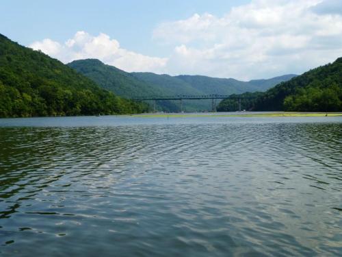 A serene lake surrounded by green mountains under a partly cloudy sky, with a bridge in the distance.
