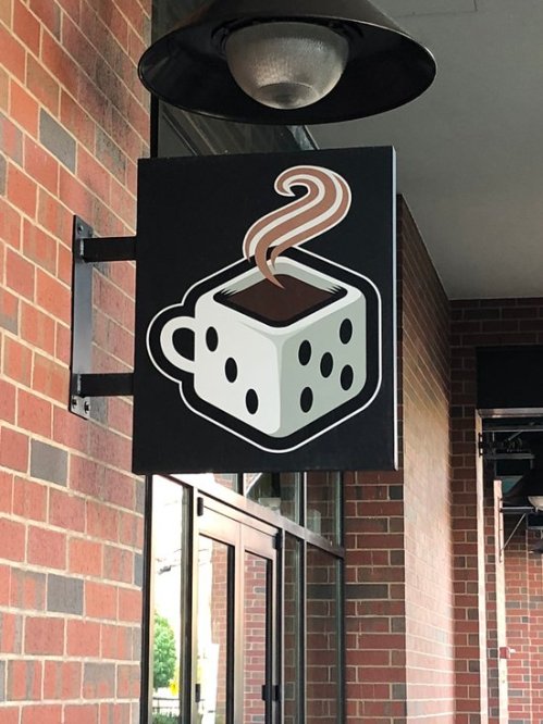 Sign featuring a stylized coffee cup with steam, designed like a die, against a brick wall.