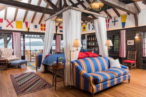 Cozy cabin interior with a four-poster bed, striped sofa, and nautical flags, overlooking a scenic lake.