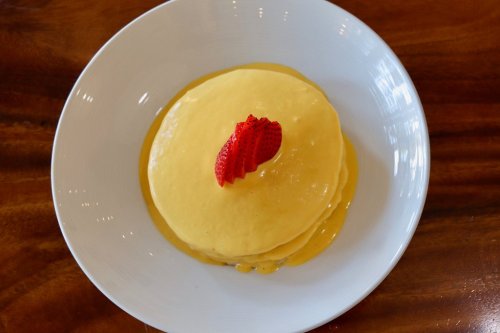 Stack of fluffy pancakes topped with a drizzle of yellow sauce and a slice of strawberry, served on a white plate.