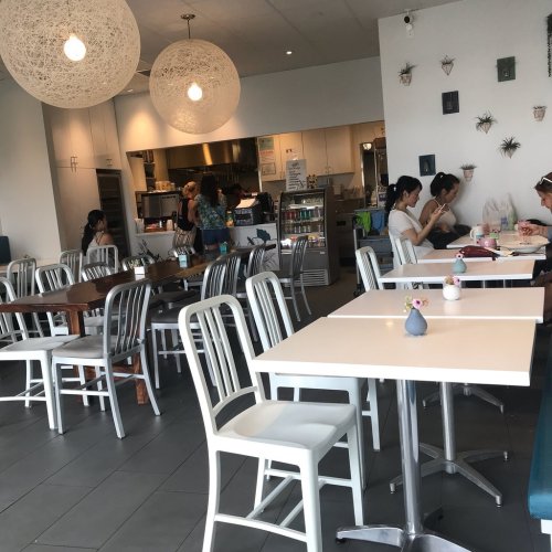 A bright café interior with white tables and chairs, featuring patrons dining and a kitchen area in the background.
