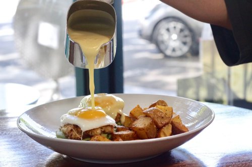 A plate of eggs benedict with a creamy sauce being poured over, accompanied by golden roasted potatoes.
