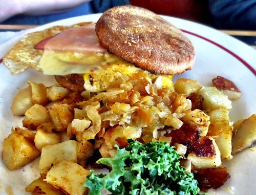 A plate of breakfast featuring a sandwich with ham and cheese, seasoned potatoes, and a side of kale.