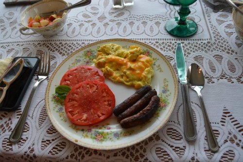 A plate with scrambled eggs, sliced tomatoes, and sausages, set on a lace tablecloth with utensils and green glasses.
