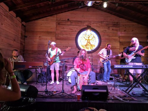 A band performs on stage in a rustic venue, featuring guitars, keyboards, and a drummer with a colorful backdrop.
