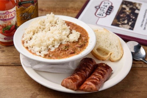 A plate of rice with a hearty stew, two sausage links, a slice of bread, and a bottle of hot sauce on the side.