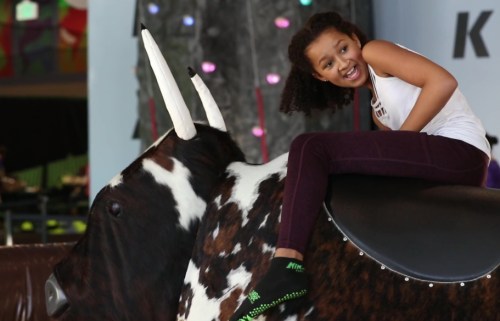 A young girl joyfully rides a mechanical bull, smiling and having fun in a colorful indoor setting.