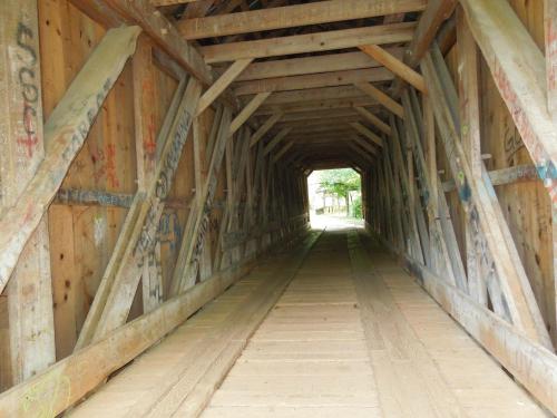 View inside a wooden covered bridge, showcasing wooden beams and a pathway leading to light at the far end.