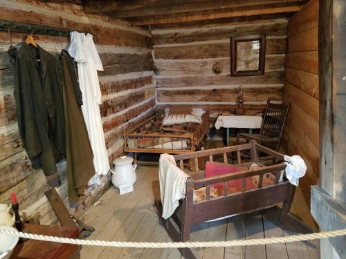 A rustic log cabin interior featuring a bed, a crib, clothing on hooks, and simple wooden furniture.