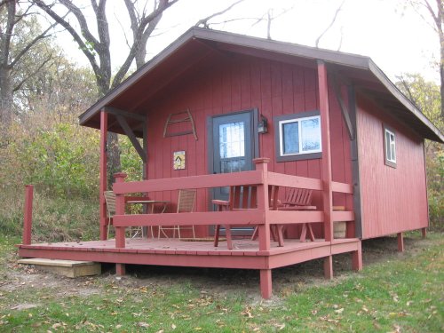 A small red cabin with a porch, surrounded by trees and greenery, featuring chairs on the deck.