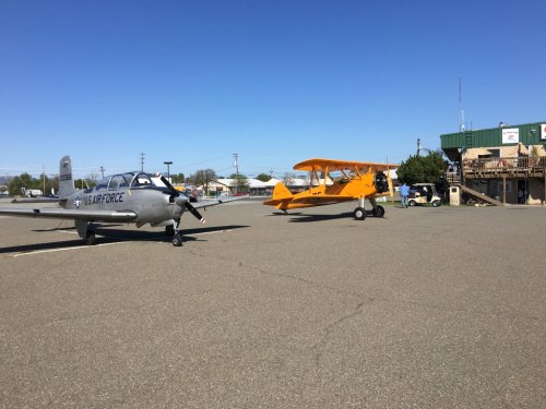 Two small airplanes parked on a tarmac under a clear blue sky, with a building in the background.