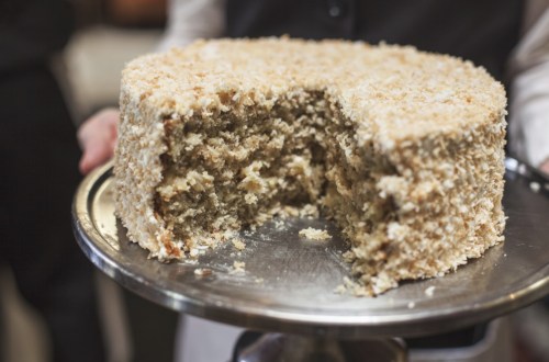 A person holds a silver tray with a coconut cake, featuring a slice cut out, revealing its moist interior.