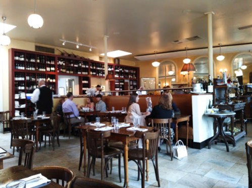 A cozy restaurant interior with wooden tables, patrons dining, and a wine display in the background.
